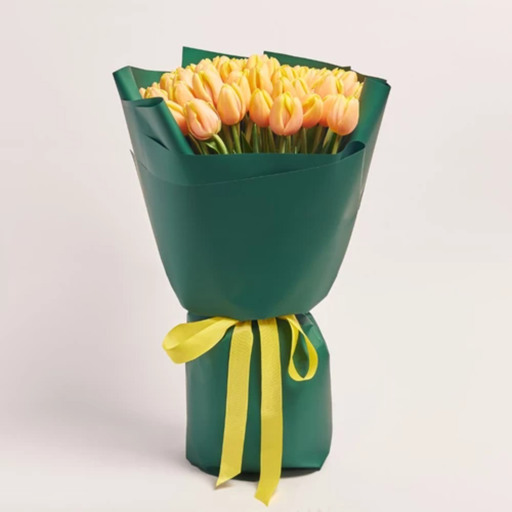 A large bouquet of peach tulips wrapped in dark green paper and tied with a yellow satin ribbon