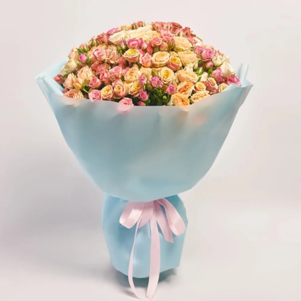 A large, dome-shaped bouquet of mixed pink and peach spray roses wrapped in light blue paper and tied with a pink satin ribbon.