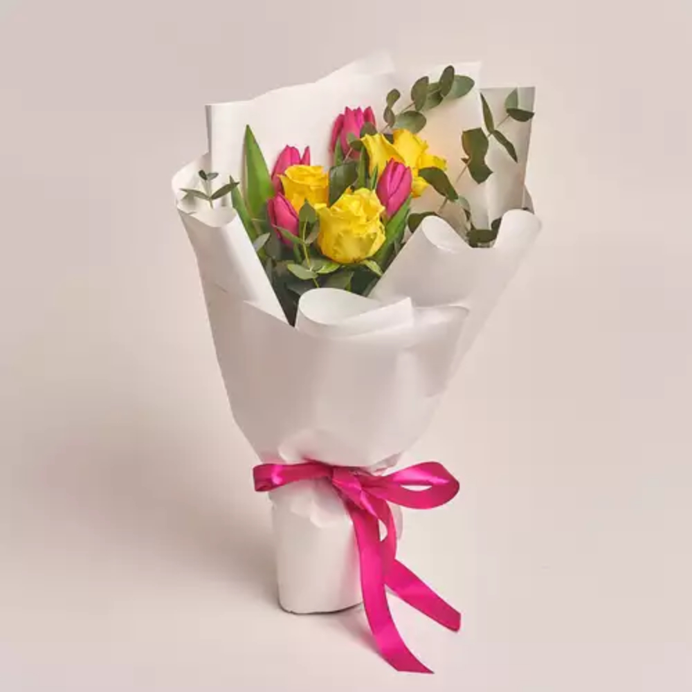 Mixed flower bouquet with yellow roses and pink tulips wrapped in white paper with a pink ribbon