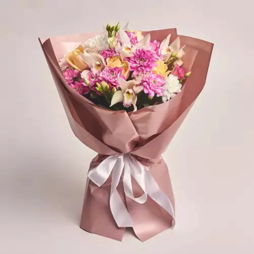 A lush bouquet of pink chrysanthemums, peach roses, white orchids, and lisianthus wrapped in dusty pink paper and tied with a white satin ribbon.