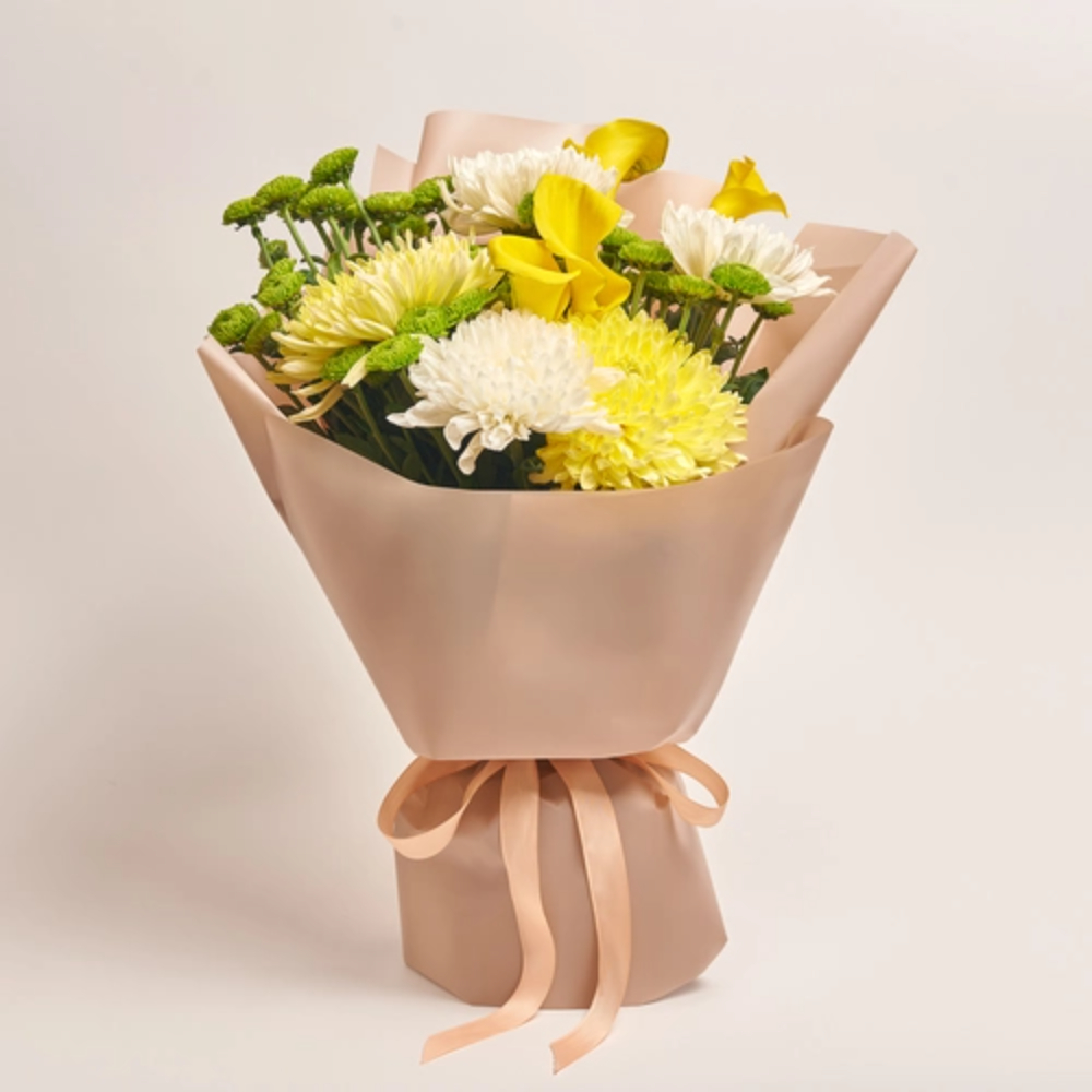 A bouquet of yellow and white chrysanthemums with yellow calla lilies and green button flowers, wrapped in beige paper and tied with a satin ribbon.