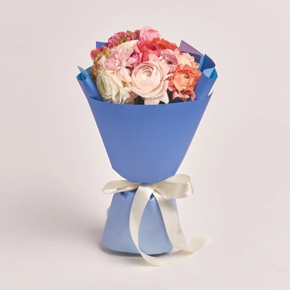 A mixed bouquet of pink, peach, and white ranunculus and roses wrapped in blue paper and tied with a white satin ribbon