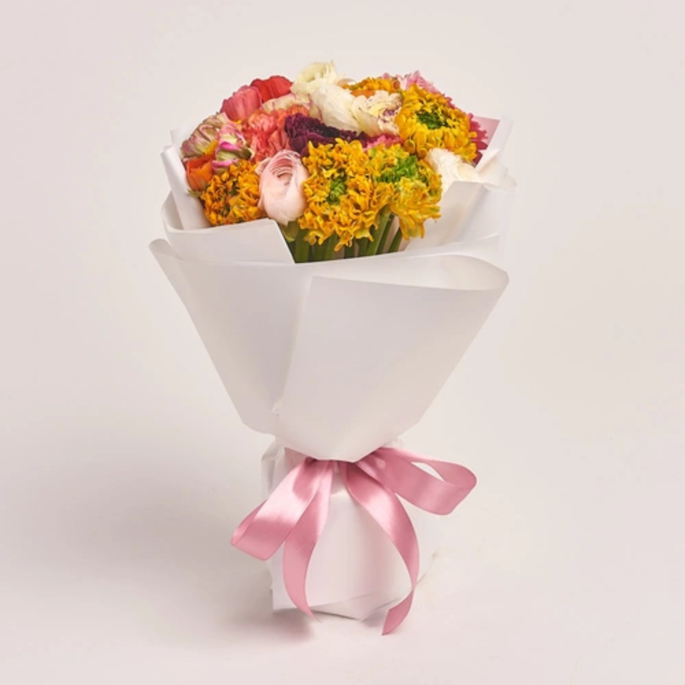 A colorful mixed bouquet of yellow, pink, orange, and white flowers wrapped in white paper and tied with a pink satin ribbon