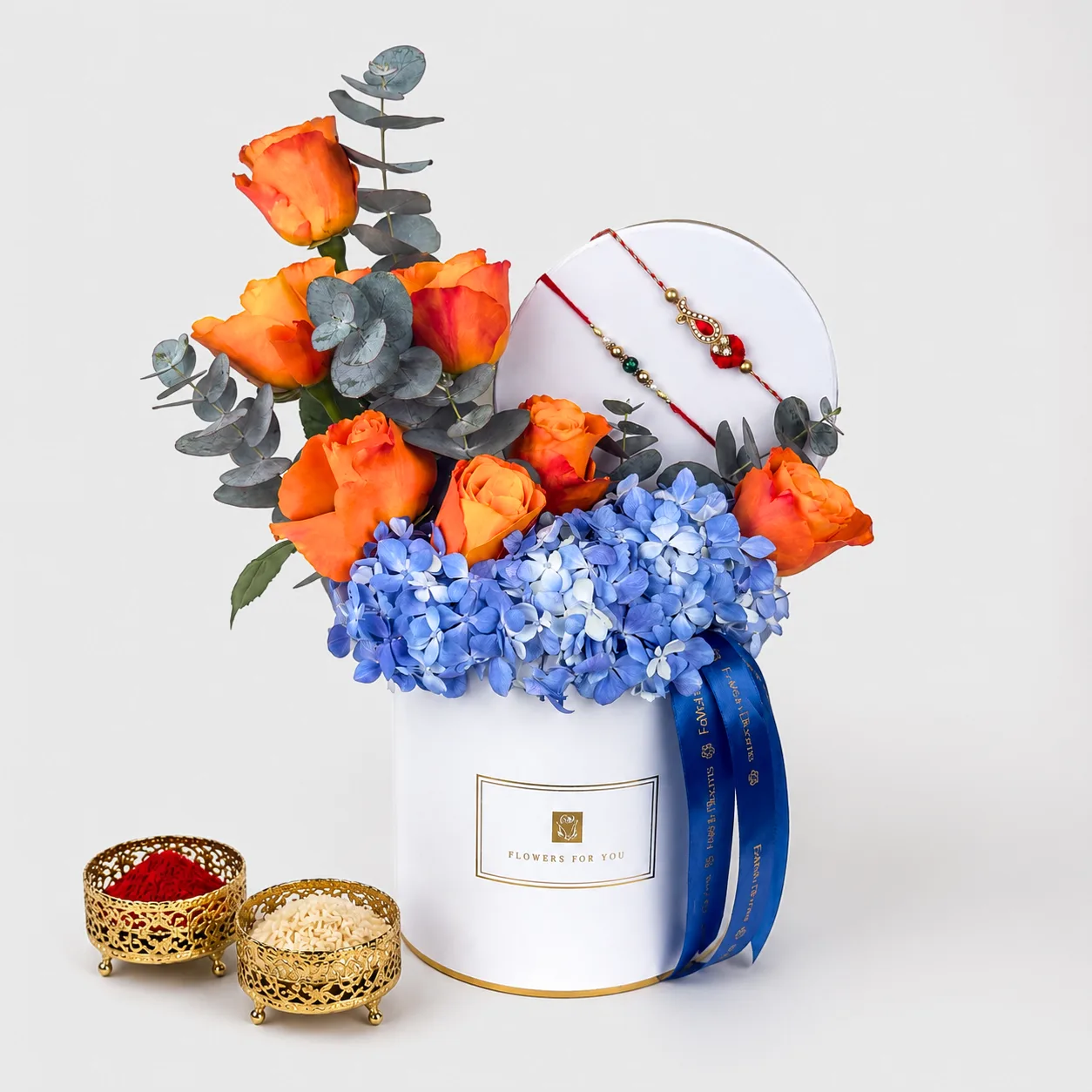 "Rakhi flower box with orange roses, blue hydrangeas, and rakhi bracelets, with kumkum and rice bowls beside it."