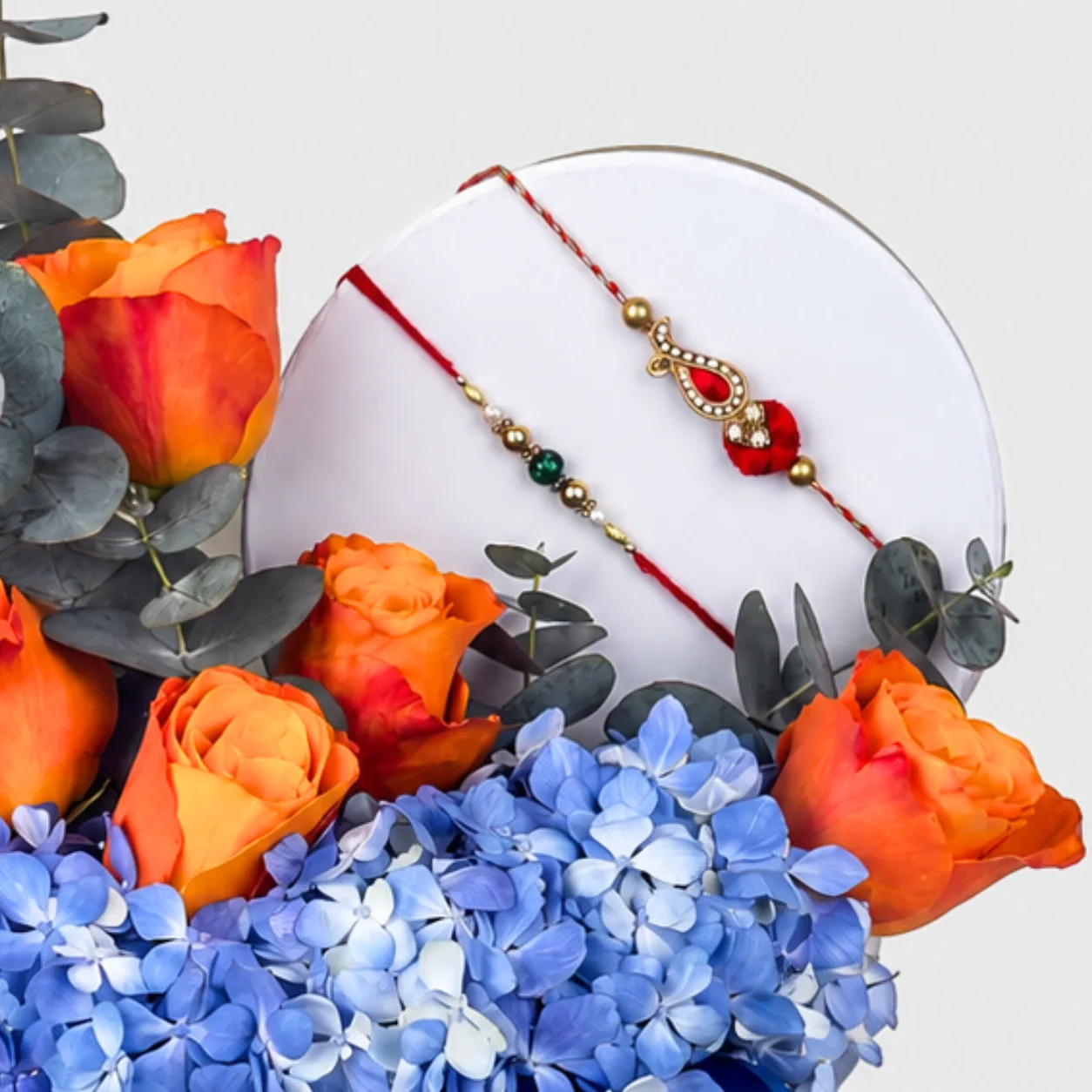 "Close-up of two rakhi bracelets on a flower box lid, surrounded by orange roses and blue hydrangeas."