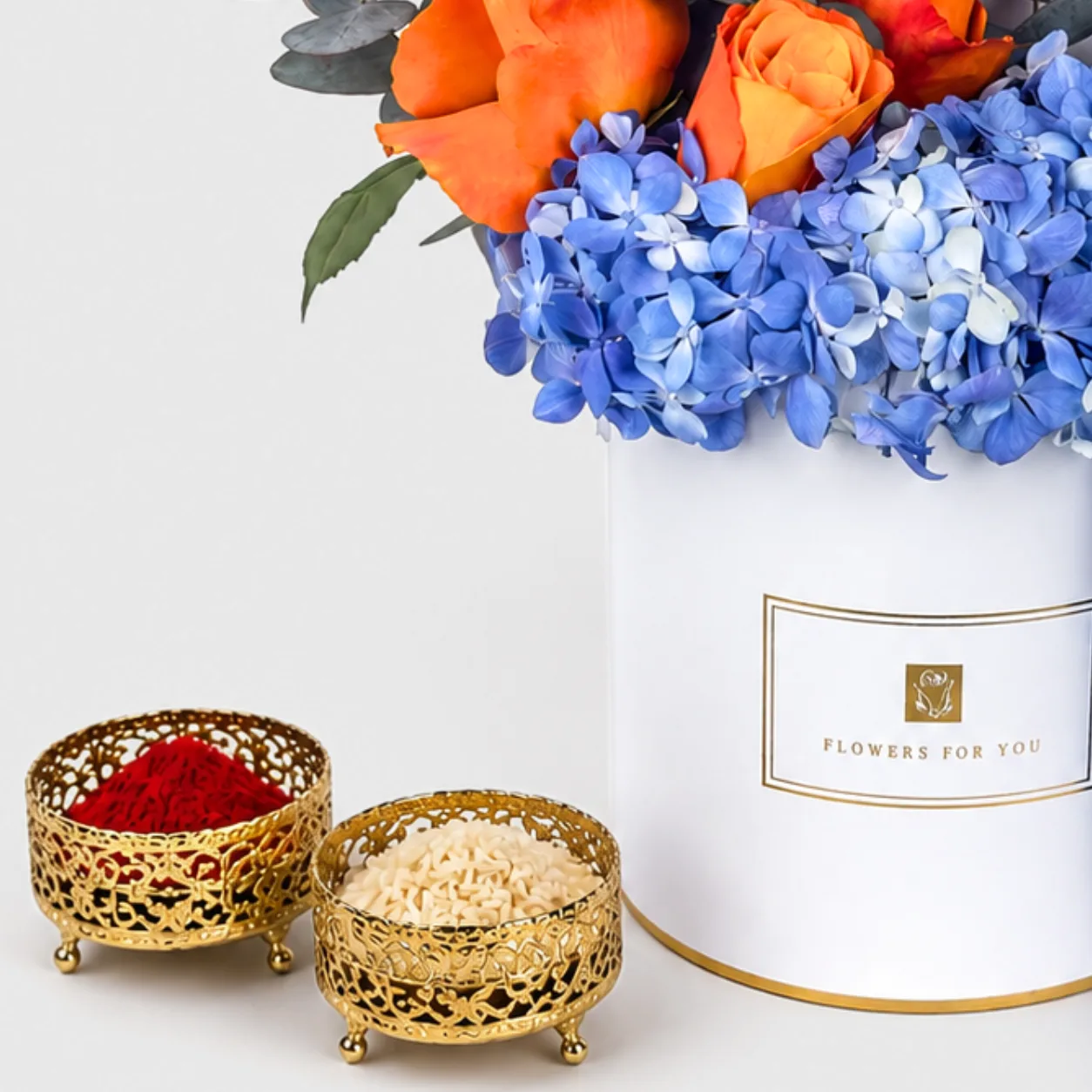 "Gold bowls of red kumkum and rice beside a white 'Flowers For You' box with orange roses and blue hydrangeas."