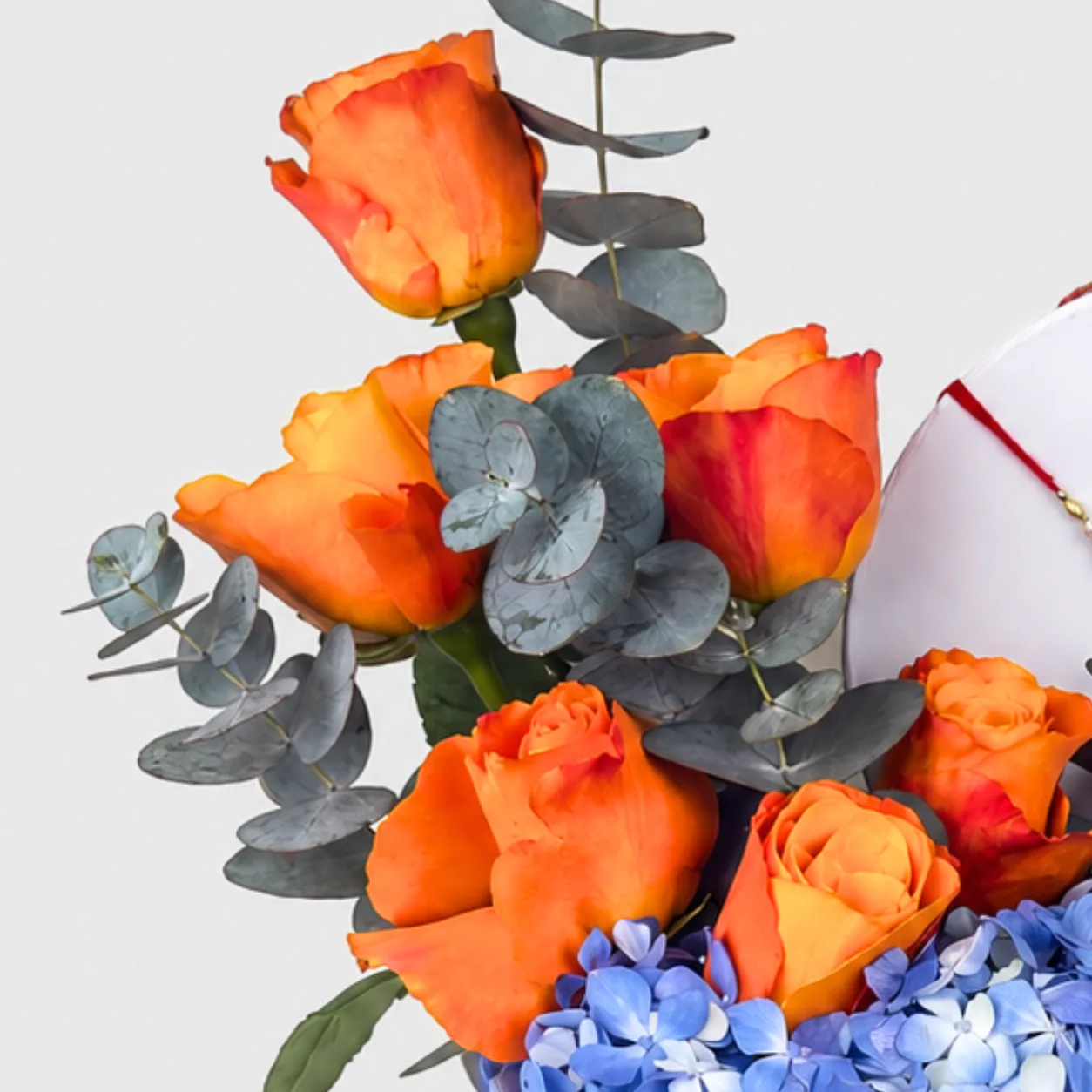 Close-up of orange roses and eucalyptus leaves with blue hydrangeas in a rakhi flower box.