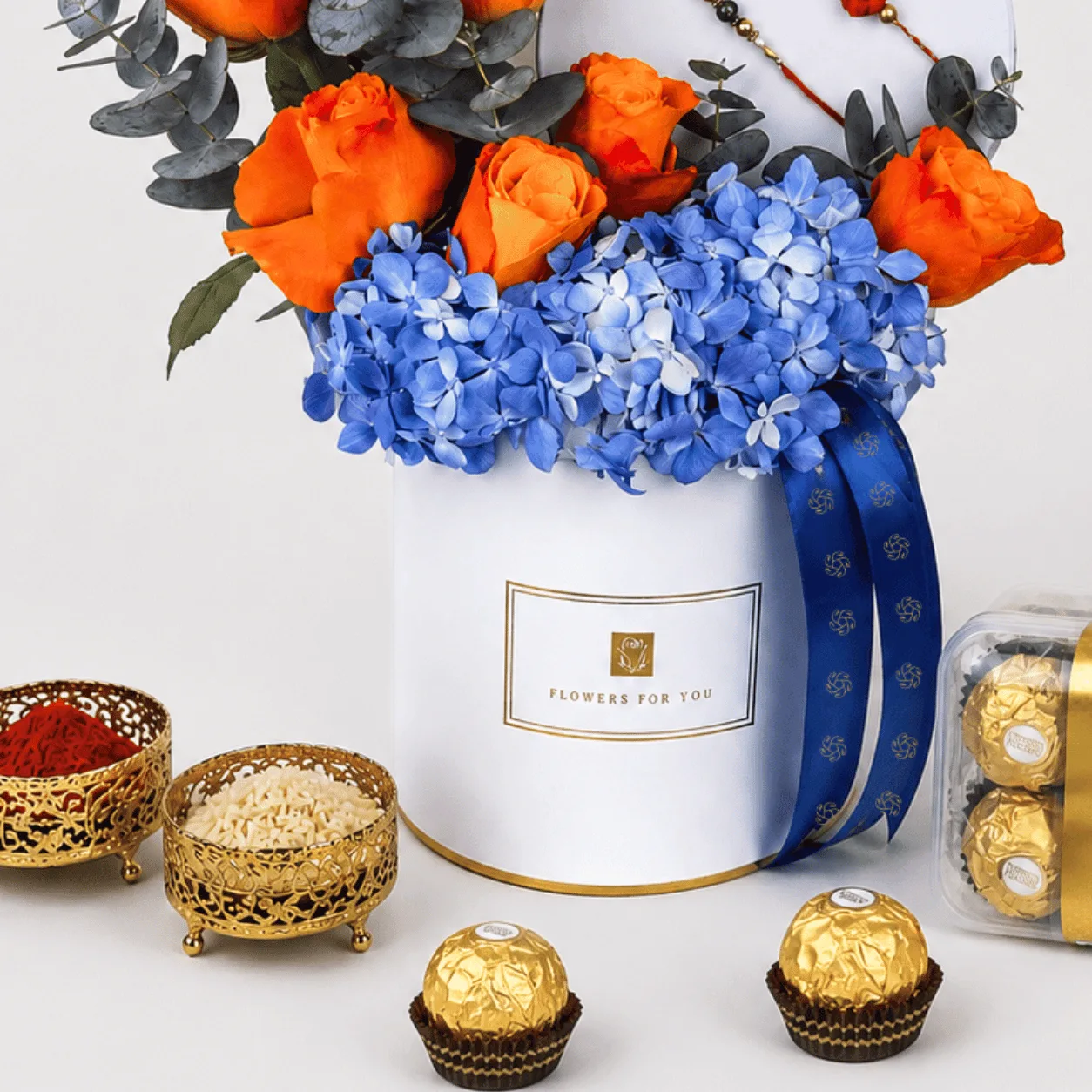 White "Flowers For You" box with orange roses and blue hydrangeas, kumkum-rice bowls, and Ferrero Rocher chocolates.