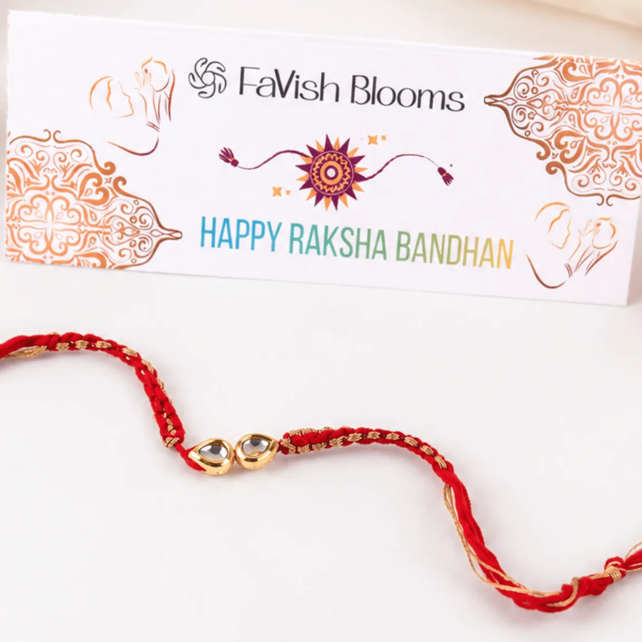 Red and gold Rakhi thread with a "Happy Raksha Bandhan" greeting card from FaVish Blooms.