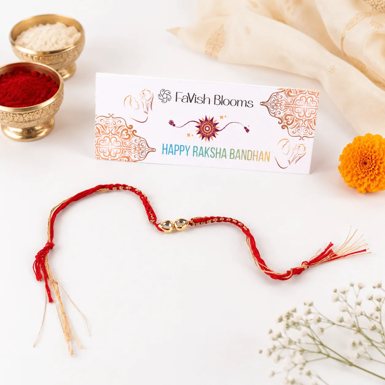 Red and gold Rakhi thread with a "Happy Raksha Bandhan" card, kumkum-rice bowls, and marigold flower.