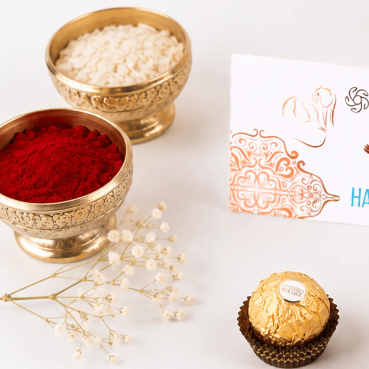 Brass bowls of kumkum and rice with a Ferrero Rocher chocolate and Raksha Bandhan card.