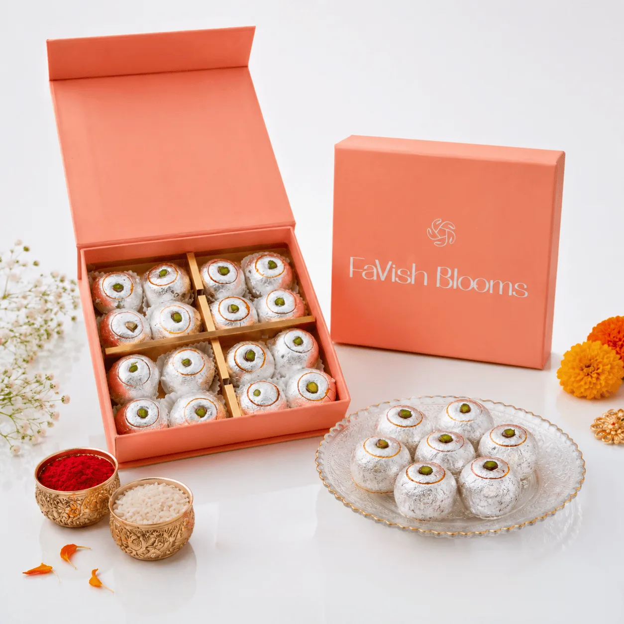Silver-foil laddus in a peach FaVish Blooms gift box, with kumkum-rice bowls and marigold flowers.
