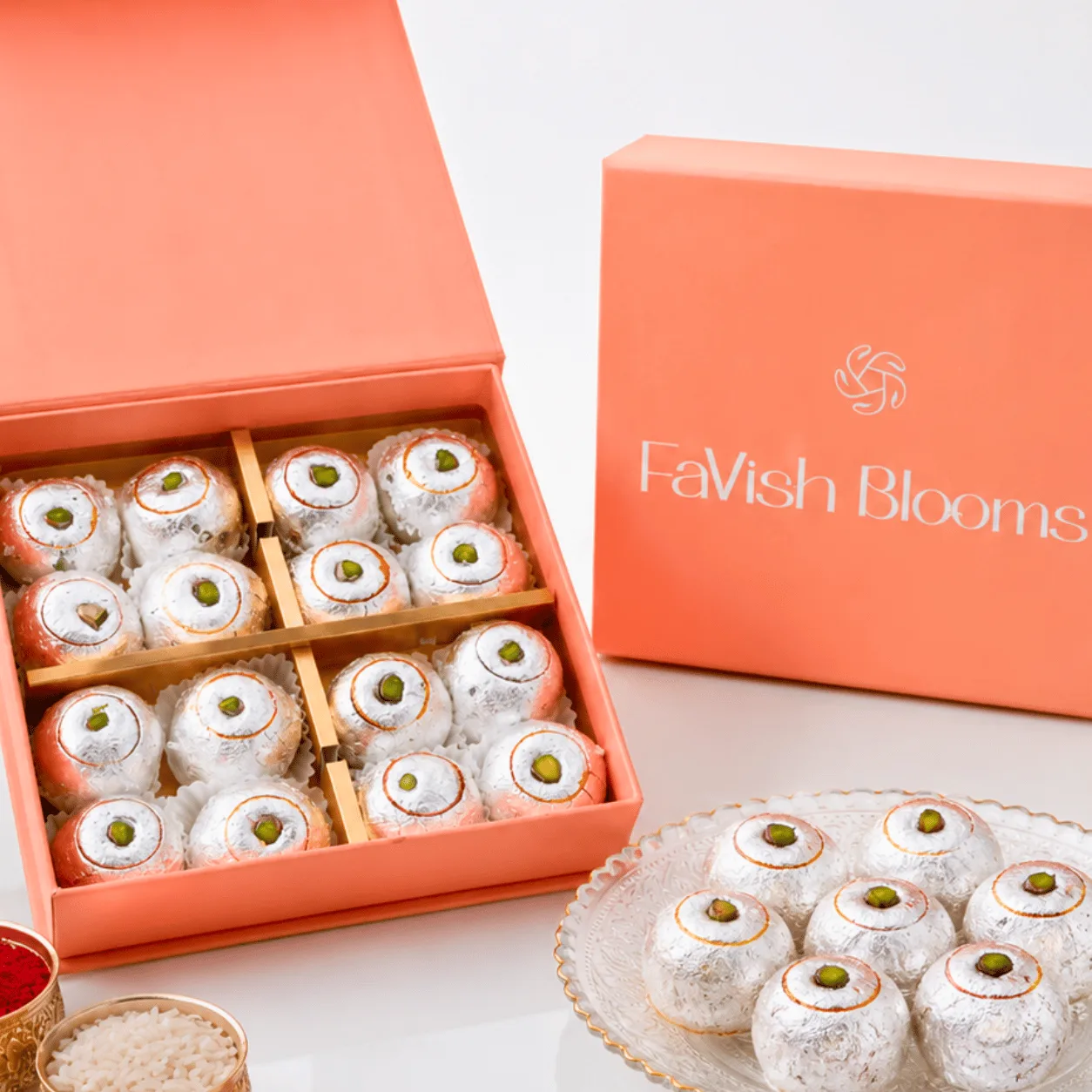 Silver-foil laddus in a peach FaVish Blooms gift box, served on a plate with kumkum-rice bowls.