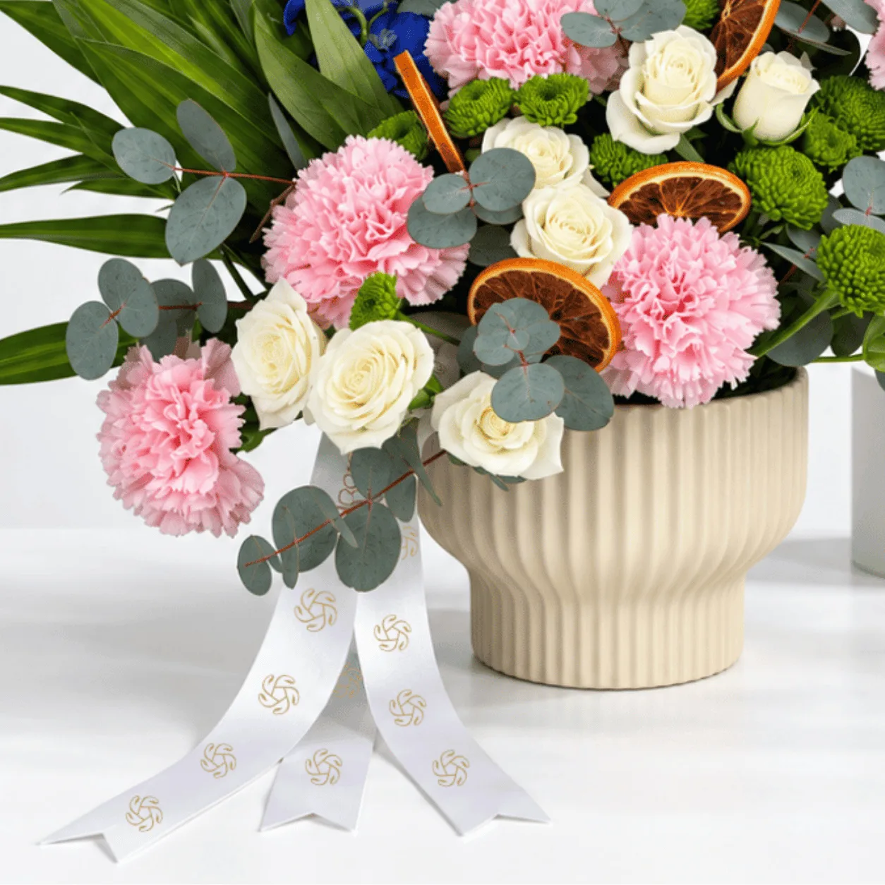 Pink carnations, white spray roses and blue delphinium arranged in a ribbed ceramic pot for Mother’s Day.