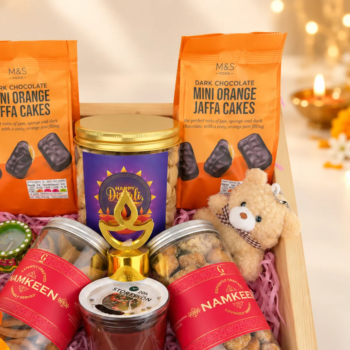 Close-up of Diwali gift hamper with chocolate Jaffa cakes, namkeen jars, teddy bear, and festive treats.