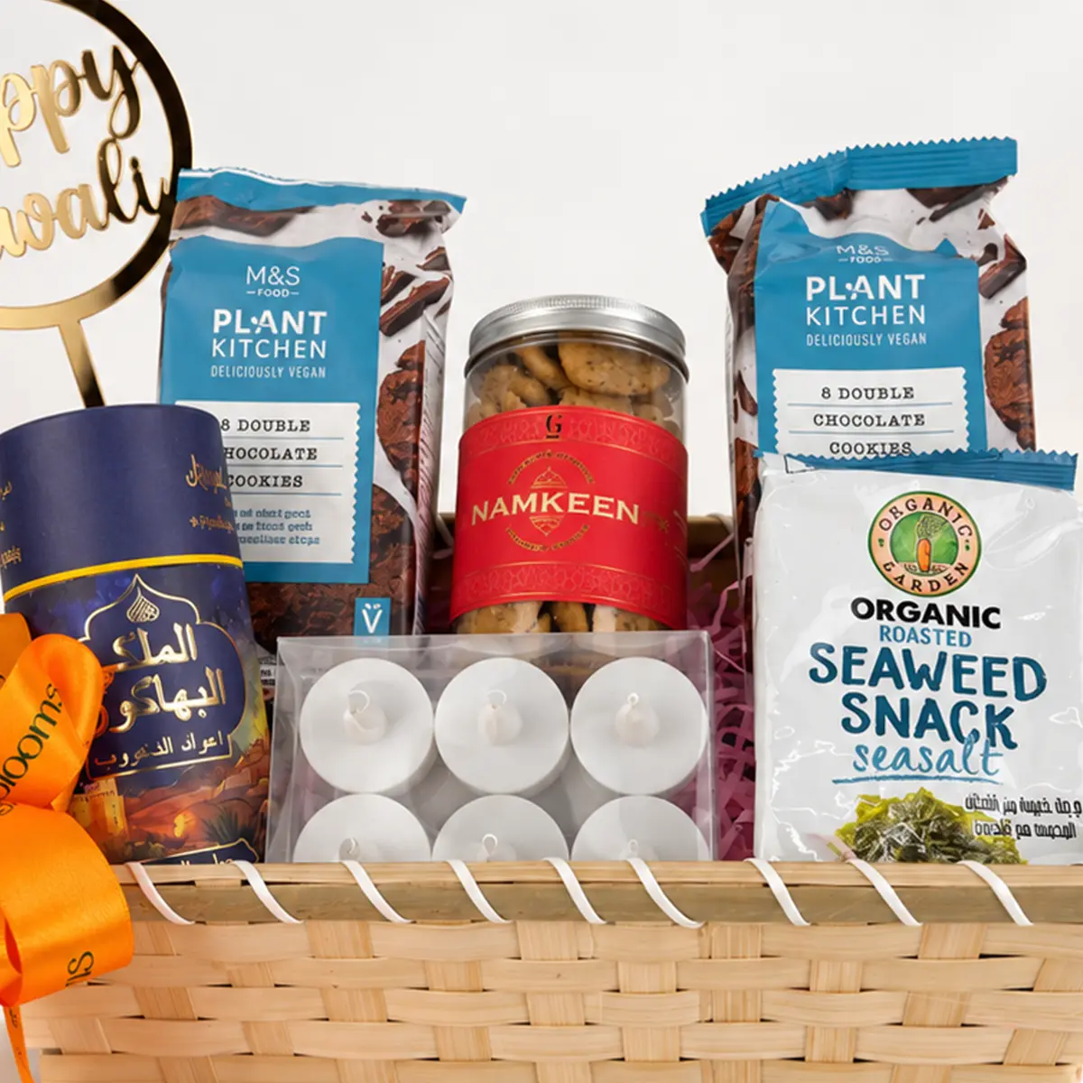 Close-up of Diwali gift hamper featuring M&S Plant Kitchen chocolate cookies, namkeen jar, organic roasted seaweed snacks, and white tealight candles in a woven basket.