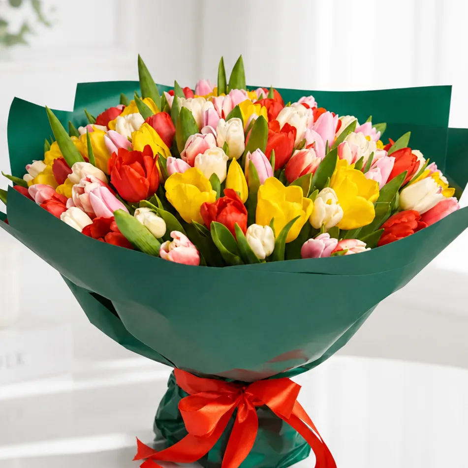 Large bouquet of mixed colorful tulips in red, yellow, pink, and white wrapped in teal paper and tied with a red ribbon on a white fur surface.