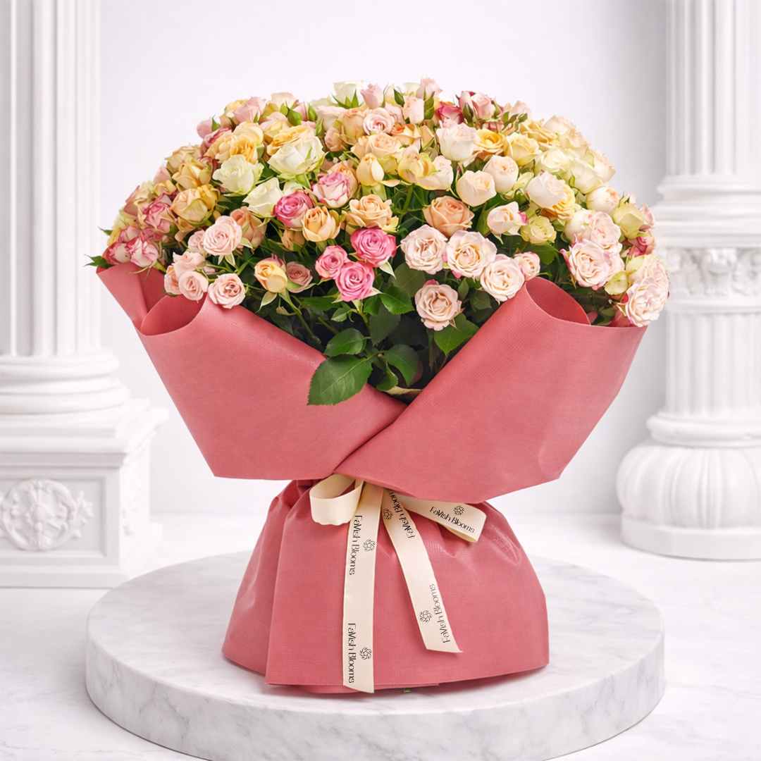 A large bouquet of mixed pastel spray roses wrapped in pink paper, tied with a cream branded ribbon.