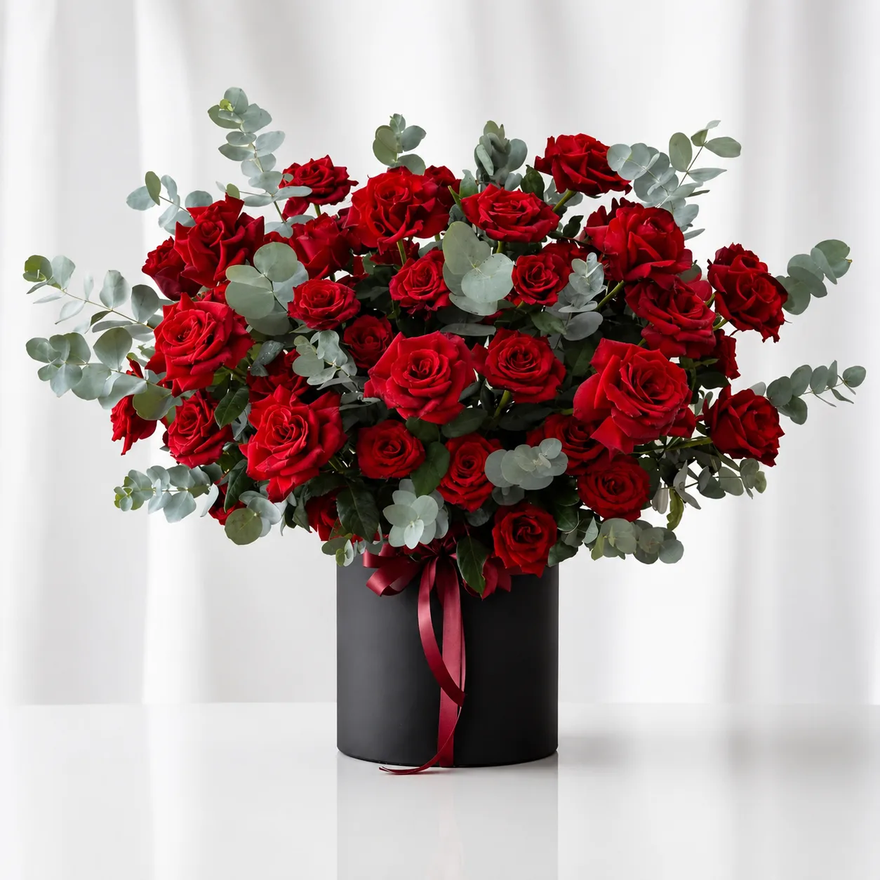 A lush arrangement of red roses and eucalyptus in a tall black cylinder box with a red ribbon, on a gold side table.