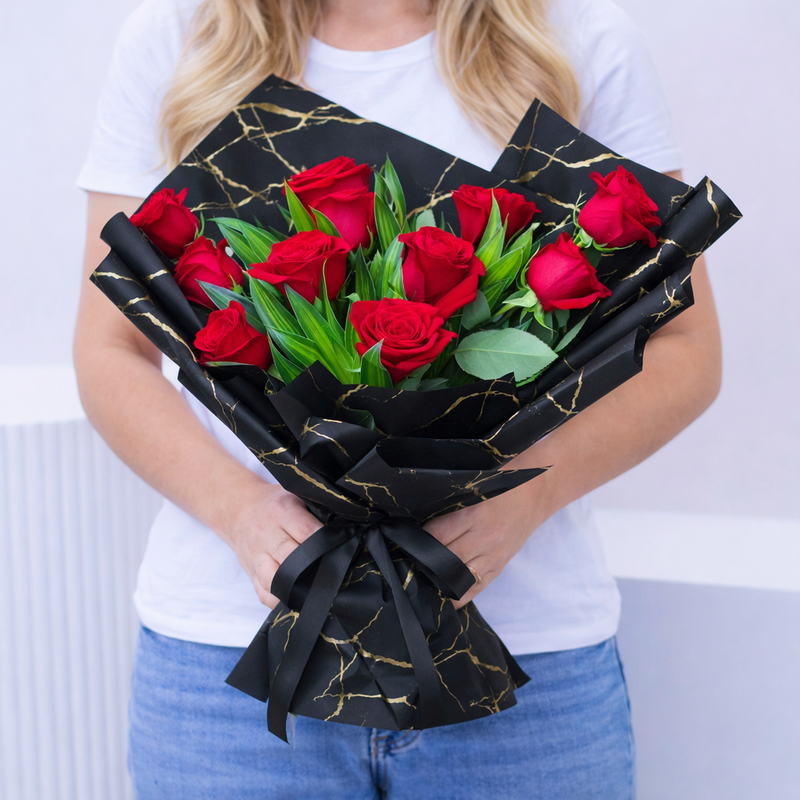 A woman holding a bouquet of red roses with greenery wrapped in black and gold marble-print paper, tied with a black ribbon.