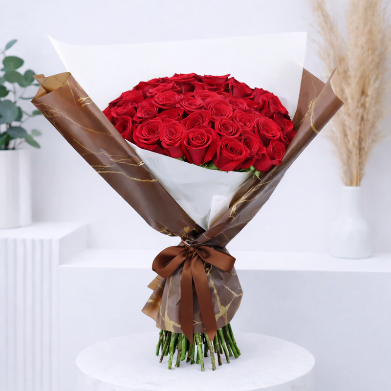 A large bouquet of red roses wrapped in brown marble-print and white paper, tied with a brown satin ribbon.