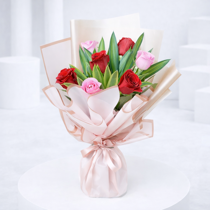 Red and pink roses with green lily leaves wrapped in blush pink paper, tied with a satin ribbon.
