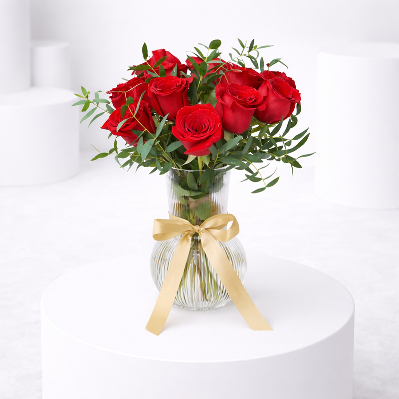 Red roses with eucalyptus in a ribbed glass vase tied with a gold ribbon bow.