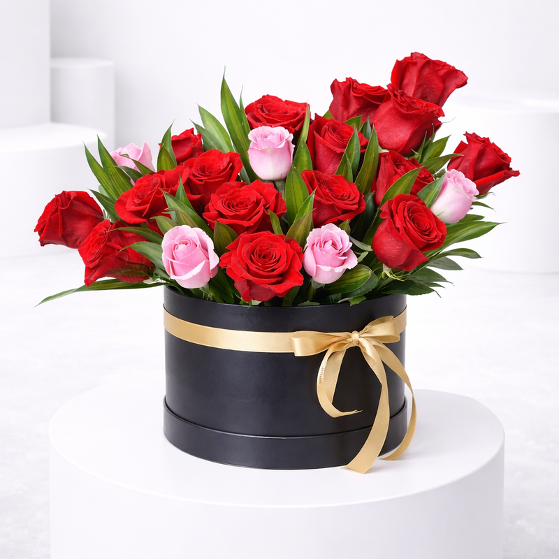 Red and pink roses with green leaves in a black round hat box with a gold ribbon bow.