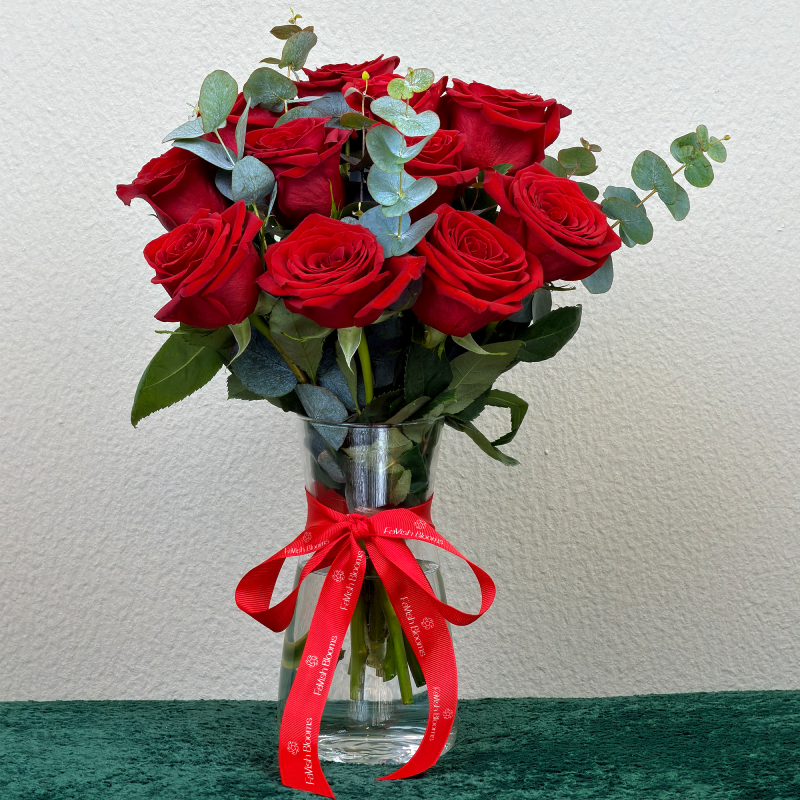 Red roses with eucalyptus in a clear glass vase tied with a red branded ribbon.