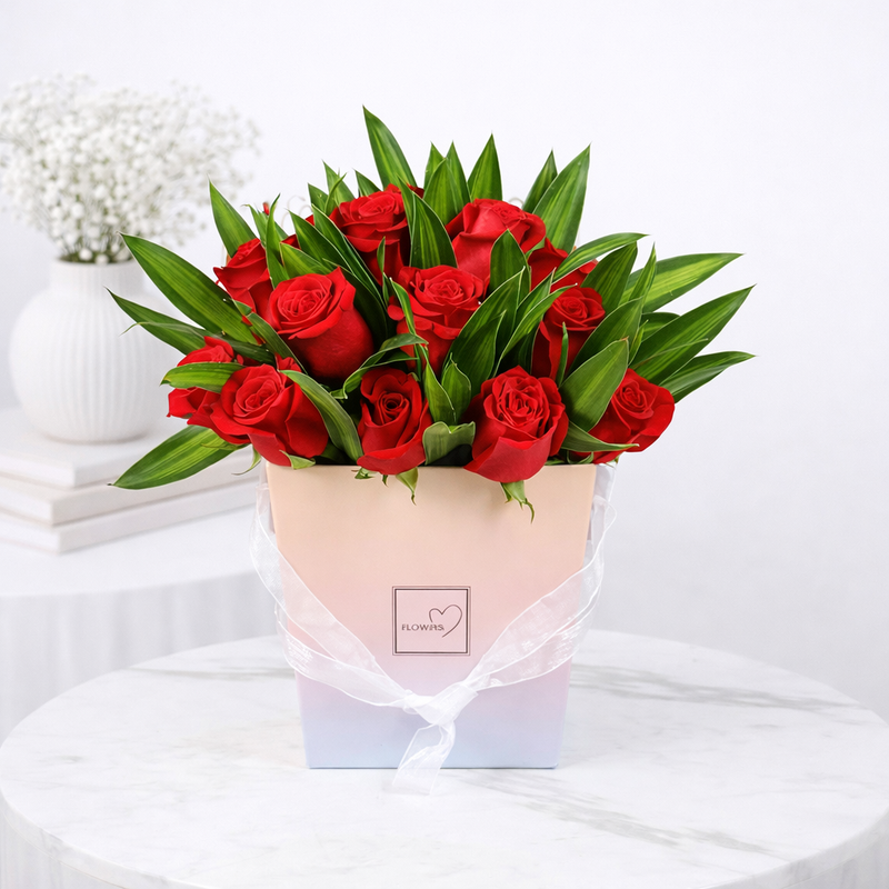 Red roses with green leaves arranged in a pastel pink gift box with a sheer white ribbon.