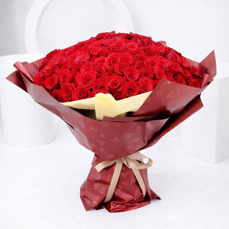 Large bouquet of red roses wrapped in burgundy heart-print paper tied with a beige ribbon.