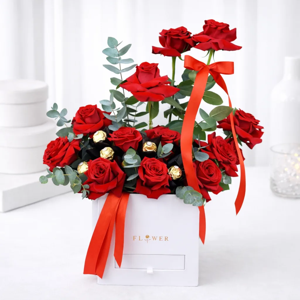 Red roses and Ferrero Rocher chocolates with eucalyptus in a white gift box tied with a red ribbon.