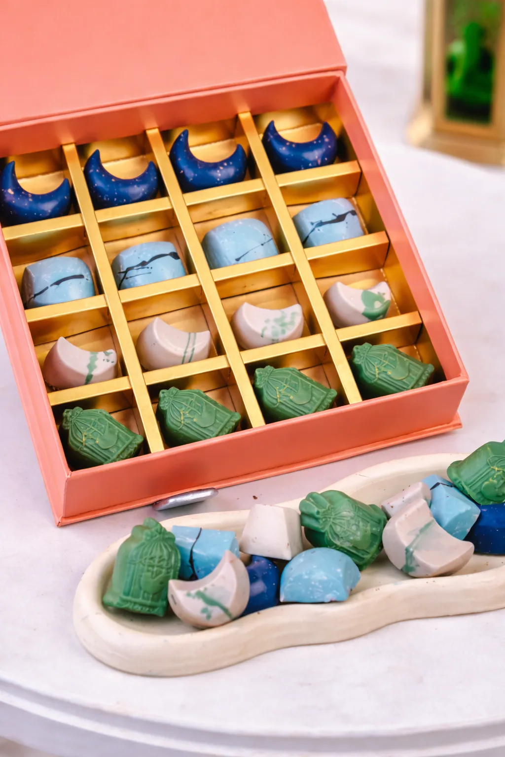 An orange-toned box contains 16 artisan chocolates arranged in a 4x4 gold grid.
