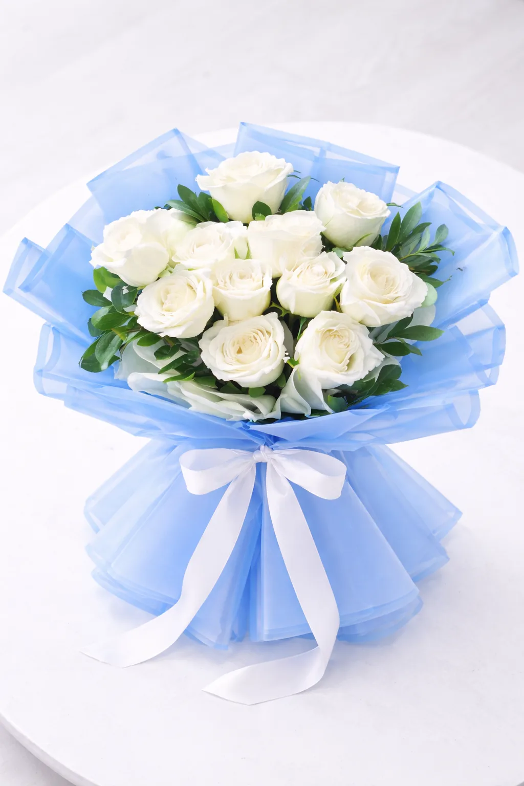 Top-down view of white roses with green leaves wrapped in blue organza and tied with a white bow.