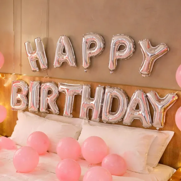 Silver 'Happy Birthday' foil letter balloons on a bedroom wall surrounded by pink balloons and fairy lights