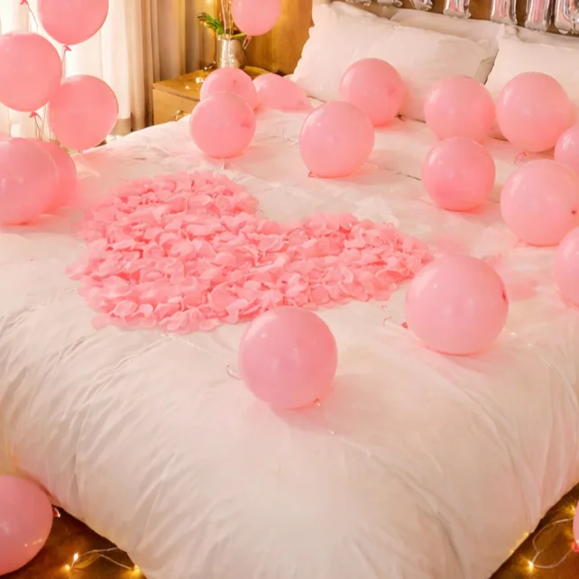 Pink rose petals arranged in a heart shape on a white bed surrounded by pink balloons and fairy lights.