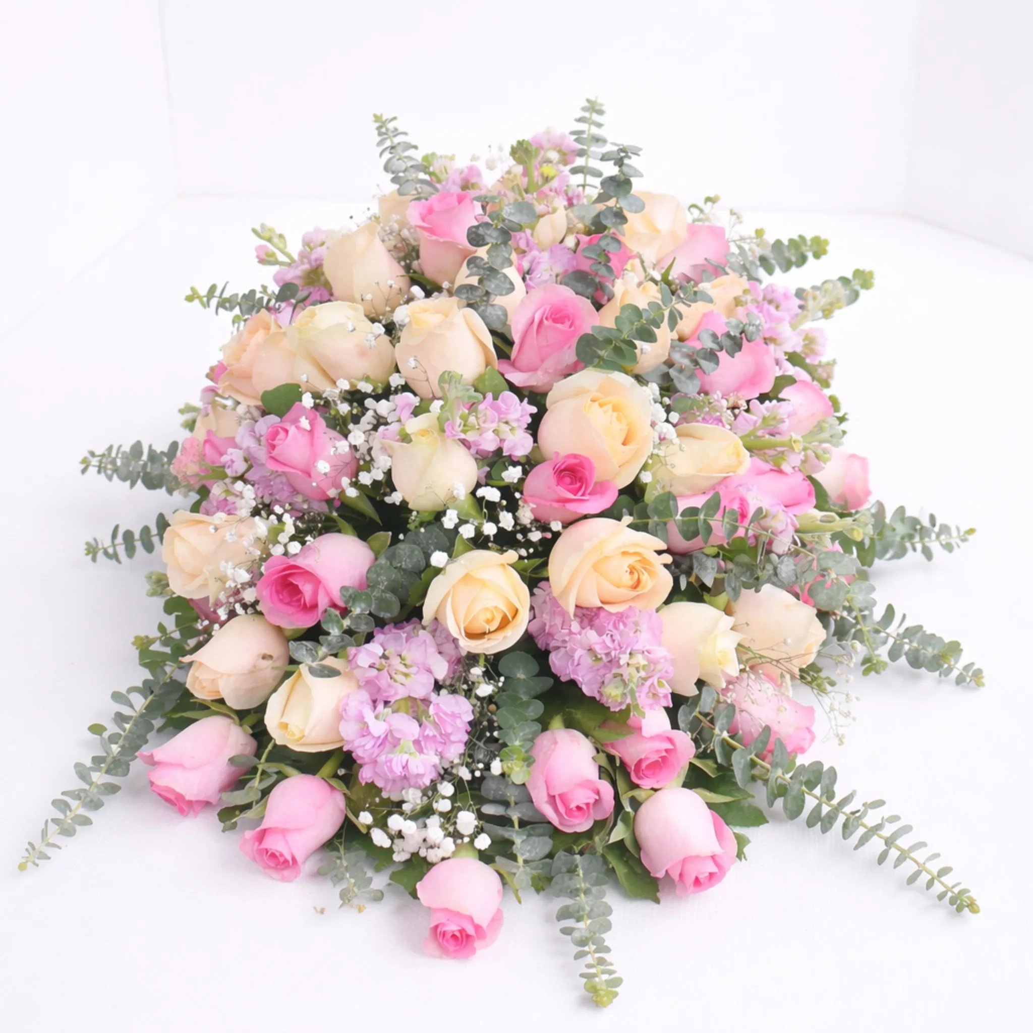 Top-down view of a lush oval floral arrangement with cream and pink roses, lavender stock, baby's breath, and trailing eucalyptus on a white surface.