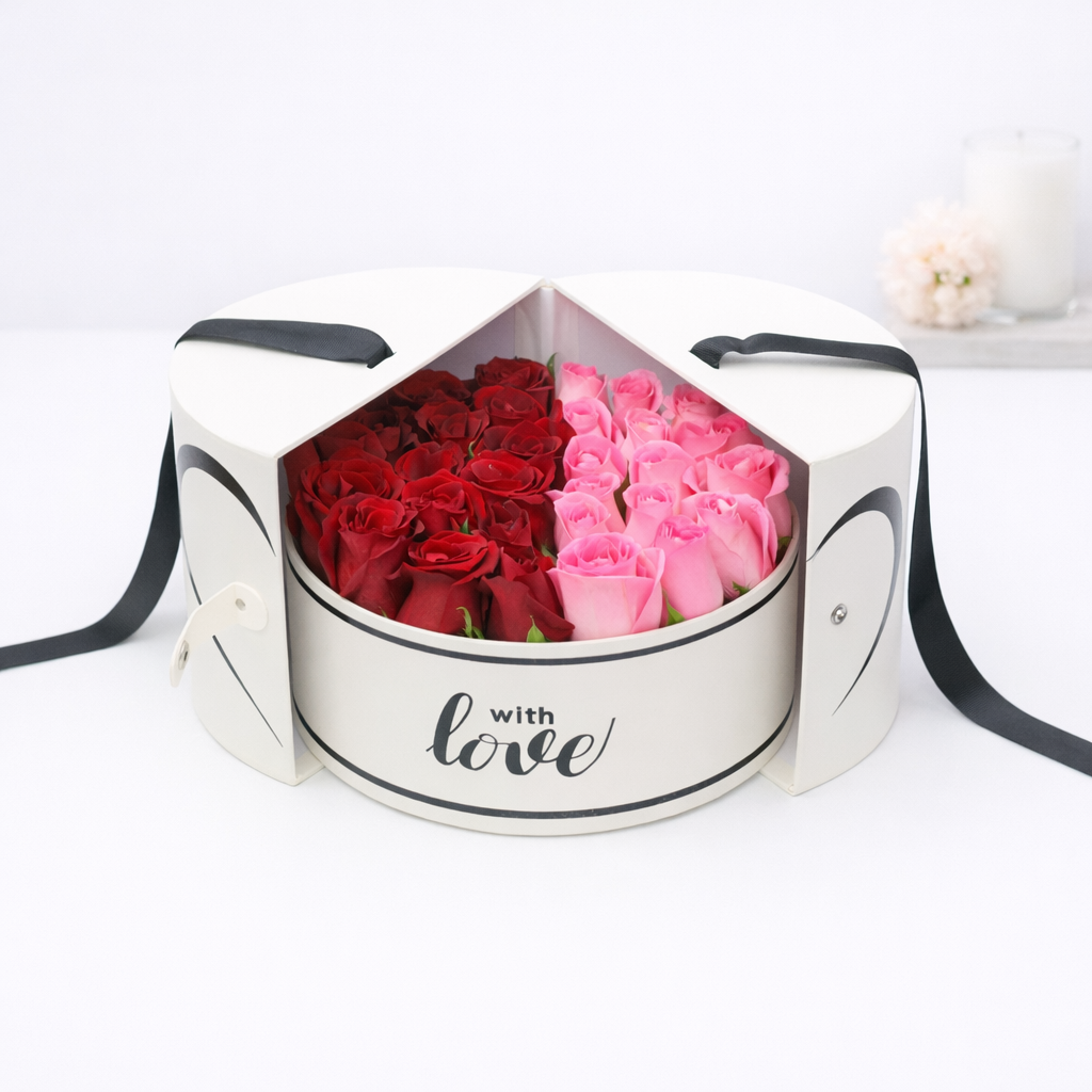 Red and pink roses arranged in a round white gift box with 'with love' script and black ribbon handles, lid open to reveal heart-shaped split.