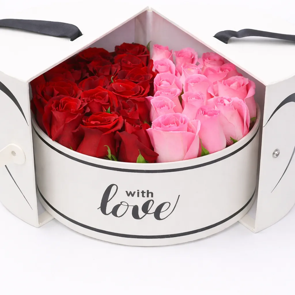 Close-up of red and pink roses filling an open round white gift box labeled 'with love,' with heart-shaped split lid and black ribbon handles.