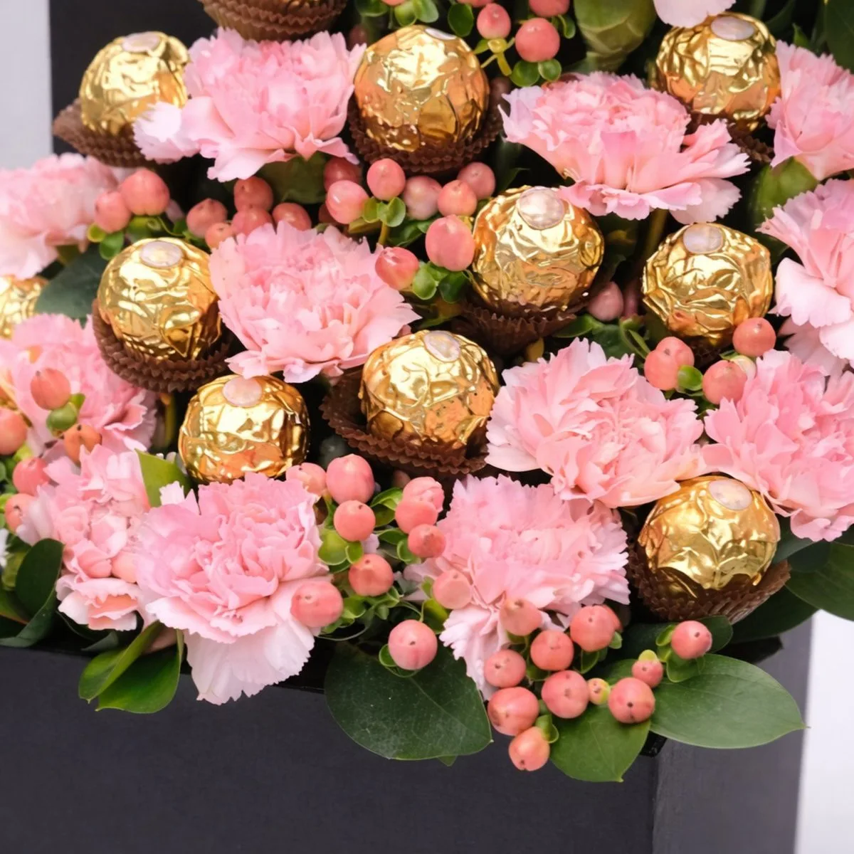 `Close-up of pink carnations, Ferrero Rocher chocolates, and peach hypericum berries in a gift arrangement.`