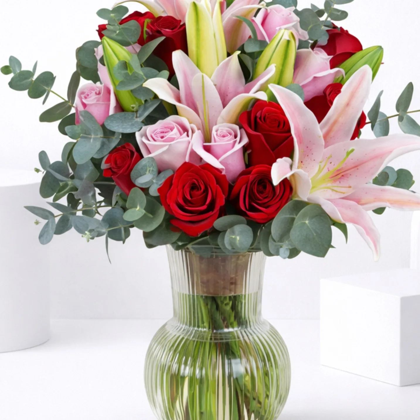A ribbed green glass vase with red roses, pink roses, and open pink lilies with eucalyptus foliage.