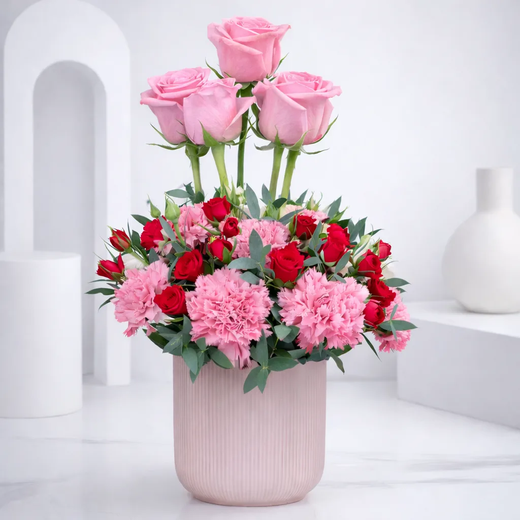 Elegant pink floral arrangement in a blush vase featuring light pink roses, pink carnations, red spray roses, and green foliage in a soft white studio setting.