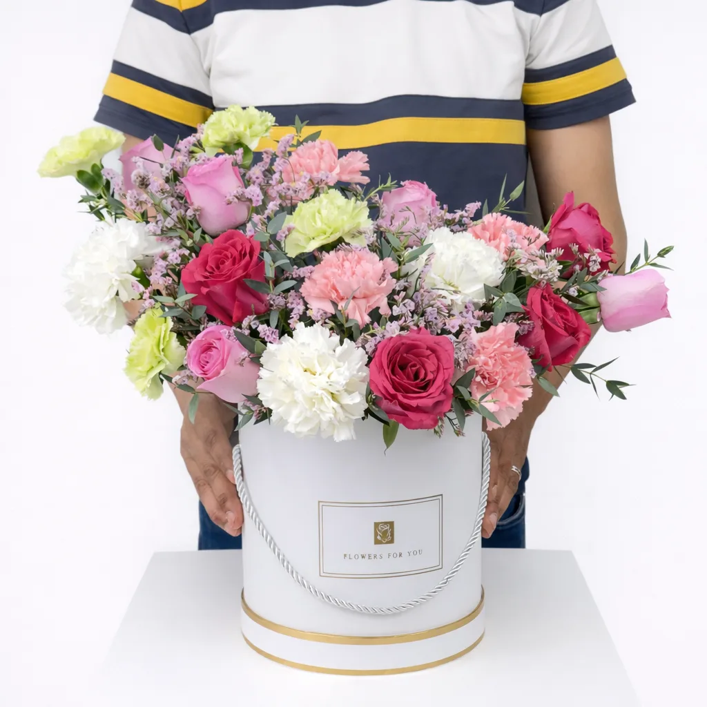 Person holding a large white round gift box with gold trim filled with red, pink, and lavender roses, white and green carnations, and purple wax flowers.