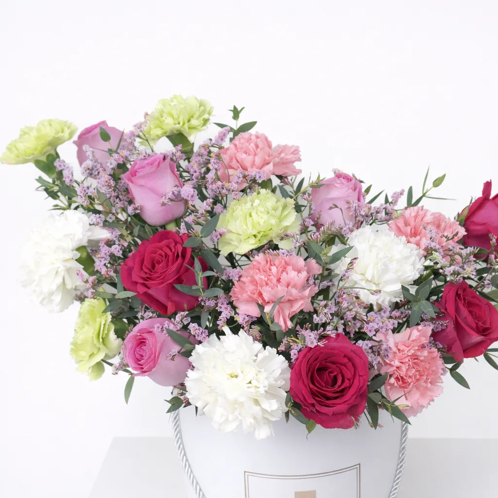 Close-up of red, pink, and lavender roses with white, pink, and green carnations and purple wax flowers in a white round gift box.