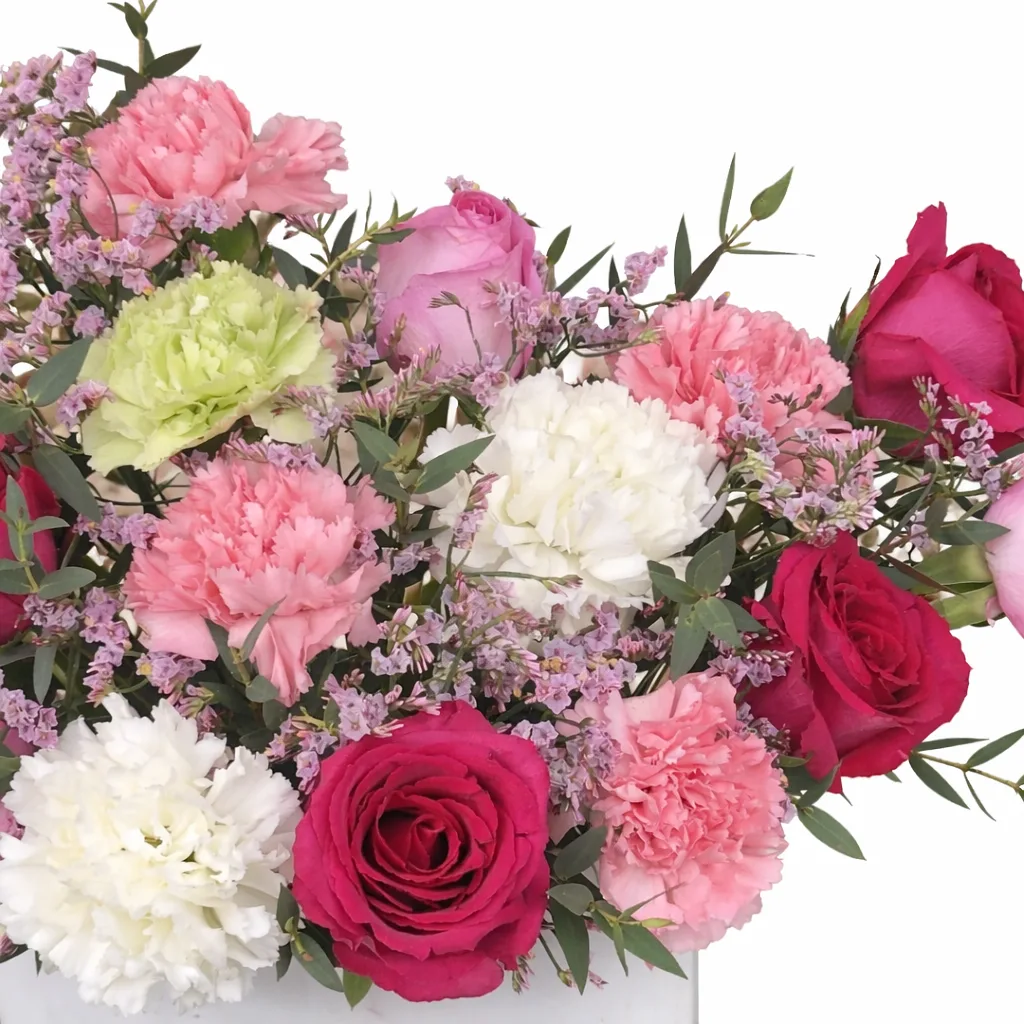 Macro shot of red, pink, and lavender roses with white, pink, and green carnations and purple wax flowers in a white box.