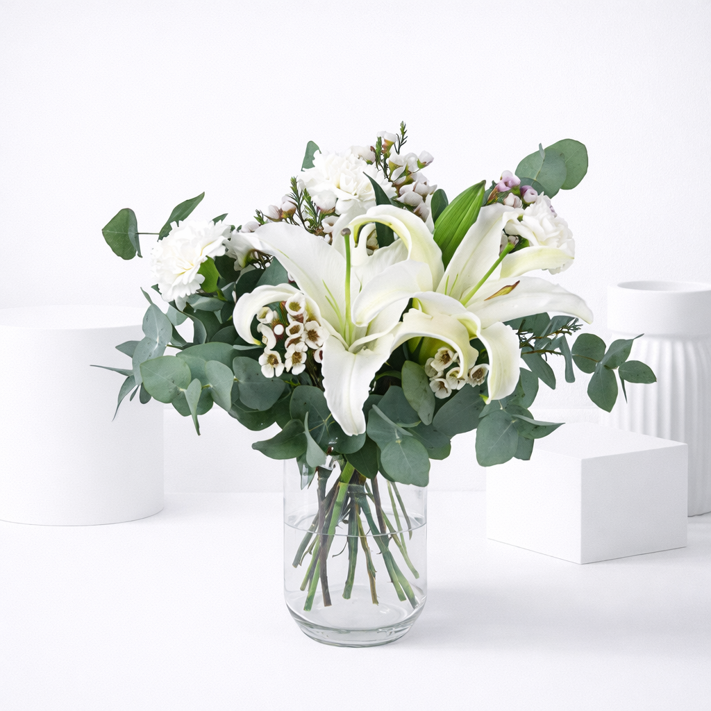 White lilies, wax flowers, and carnations with eucalyptus arranged in a clear glass vase.