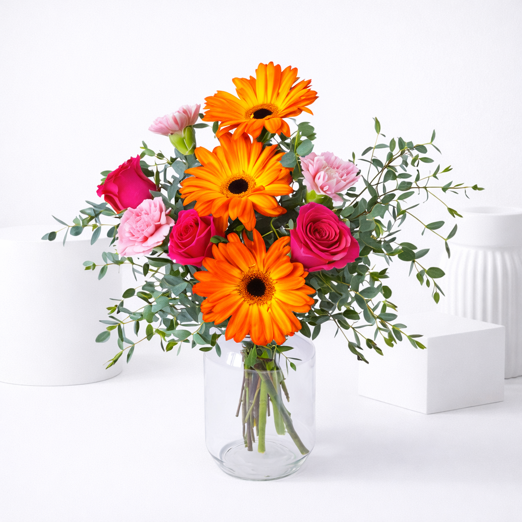 Orange gerbera daisies, hot pink roses, and pink carnations with eucalyptus in a clear glass vase.