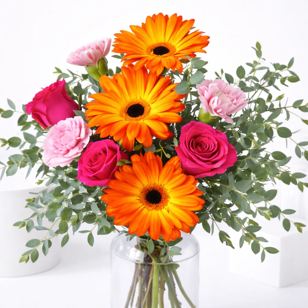 Close-up of orange gerbera daisies, hot pink roses, and pink carnations with eucalyptus in a clear glass vase.