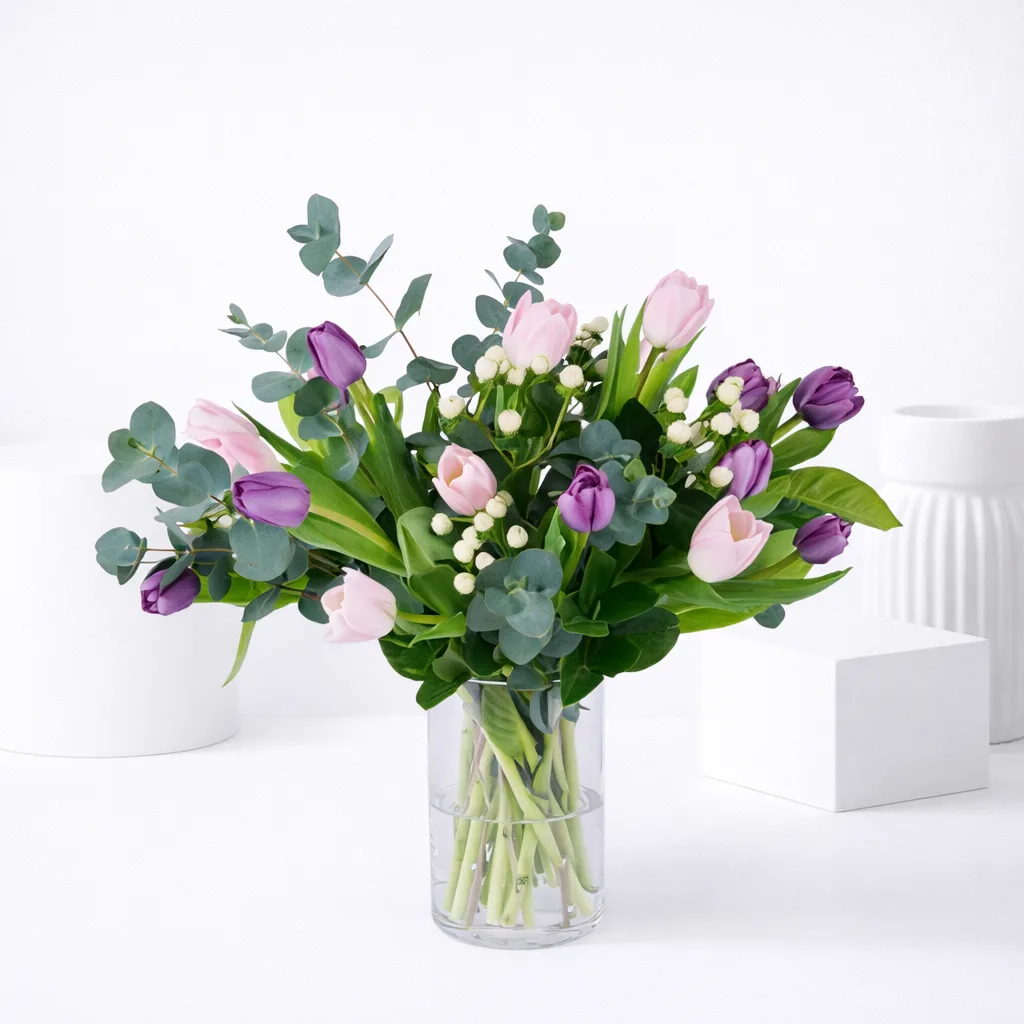 Pink and purple tulips with white berry stems and eucalyptus arranged in a clear glass vase.