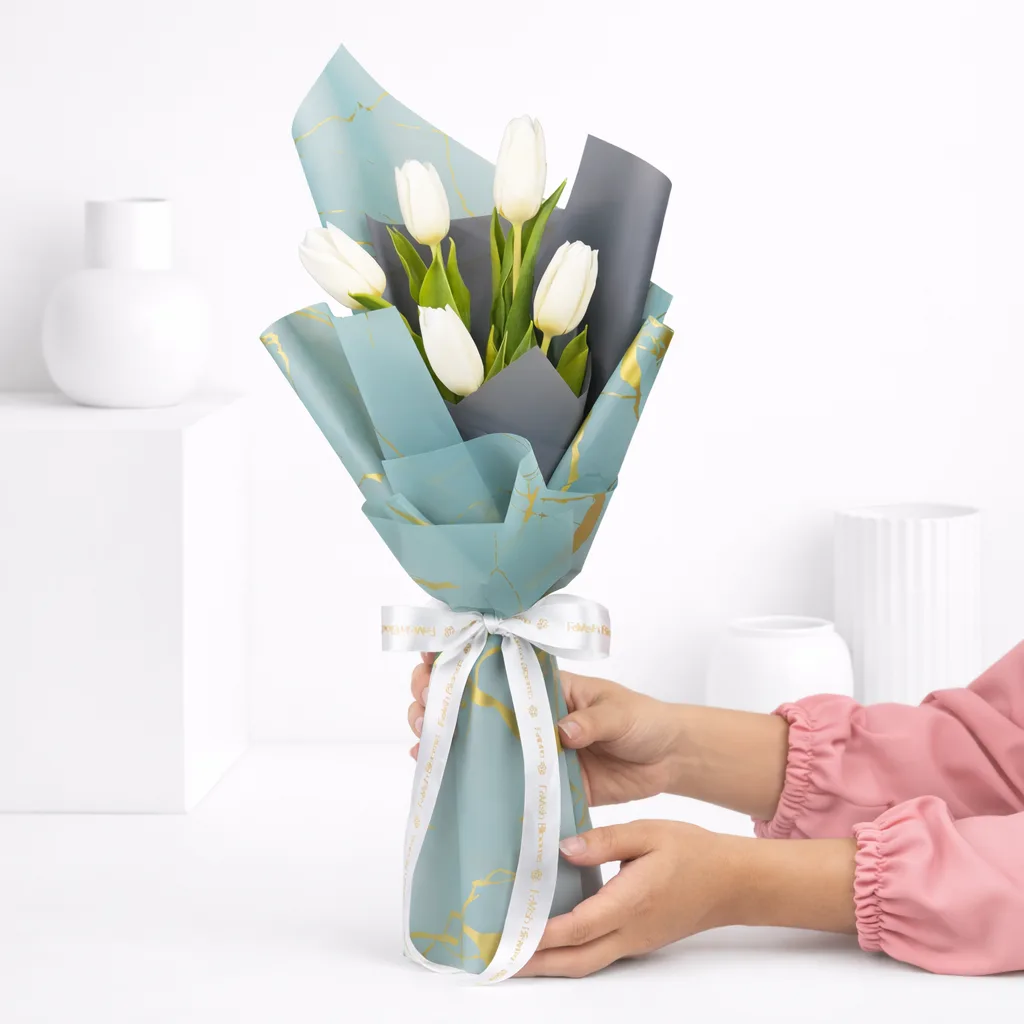 Hands holding a white tulip bouquet wrapped in sage green marble paper with a white ribbon bow.